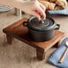 A hand using a chestnut finish display stand to place a lid on a small black Valor cast iron pot.