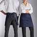 A man and woman wearing navy blue half bistro aprons with pockets.