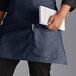 A woman wearing a navy blue half bistro apron holding a white notebook and a pen.