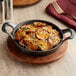 A Valor pre-seasoned cast iron casserole dish with potatoes, onions, cheese, and rosemary on a wooden table with a fork on a napkin.