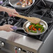 A person cooking vegetables in a Vollrath stainless steel fry pan on a stove top.