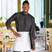 A man wearing a black shirt and white Choice half bistro apron standing in a professional kitchen.