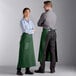 A man and woman wearing Choice hunter green bistro aprons standing in a professional kitchen.