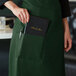 A woman wearing a Choice hunter green standard bistro apron with a black book and pen in the pocket.