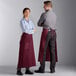 A man and woman wearing burgundy bistro aprons with pockets.