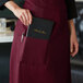 A woman wearing a burgundy standard bistro apron with a pocket and putting a black book in it.