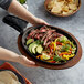 A person holding a Valor cast iron fajita skillet with meat and vegetables over a plate of food.
