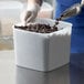 A hand in a white glove using a scoop to pour chocolate chips into a white Carlisle food storage container.
