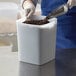 A person pouring coffee beans from a metal scoop into a white square Carlisle food storage container.