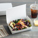 A salad in a Fold-Pak white paper take-out box with a drink and a plastic fork on a napkin.