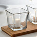 Two square Anchor Hocking glass votive holders with candles on a wooden tray.