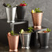 Four Acopa copper mint julep cups with ice, lemon, and mint leaves on a table with other fruit and metal cups.