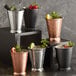 A group of Acopa Alchemy hammered copper mint julep cups with fruit and leaves on a table with a silver bucket of ice and fruit.