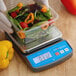 A hand holding a plastic container of vegetables on an AvaWeigh digital portion scale.