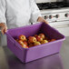 A woman in a chef's uniform holding a purple Vollrath bus tub full of red apples.