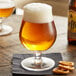 A close up of a glass of Arcoroc Belgian beer with foam, next to a bowl of pretzels.
