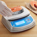 A hand wearing gloves uses an AvaWeigh digital portion scale to weigh a piece of salmon.