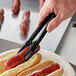 A hand using Visions black tongs to add a hot dog to a bun.