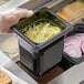 A hand in a plastic glove holding a Vigor 1/6 size black polycarbonate food pan filled with lettuce.
