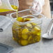A gloved hand pouring liquid from a plastic container into a clear plastic food pan filled with pickles.