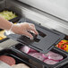 A person wearing plastic gloves putting a Vigor black polycarbonate food pan lid on a plastic tray of food.