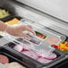 A gloved hand using a Vigor clear polycarbonate food pan lid with a handle on a plastic tray of food.