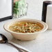 A tan Acopa Foundations melamine bowl filled with soup, noodles, and meatballs sits on a table next to a spoon.