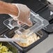 A person wearing a glove uses a Vigor 1/6 size clear plastic food pan lid with a notch and handle to cover a container of food.
