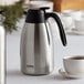 A Thermos stainless steel coffee carafe on a table with a white cup filled with brown liquid.