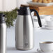 A silver and black Thermos FN371 stainless steel coffee carafe on a table with a cup of coffee.