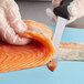 A person in a plastic glove using a Mercer Culinary semi-flexible fillet knife to cut a piece of salmon.