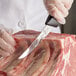 A gloved hand uses a Mercer Culinary curved boning knife to cut meat.