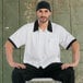 A man wearing a white Uncommon Chef cook shirt with black trim sitting on a stool.