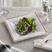 A Visions white plastic square plate with rose gold and copper bands holding a salad on a table.