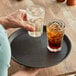 A person holding a Choice black round non-skid serving tray with a drink and a glass of ice.
