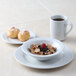 A Tuxton bright white china bowl filled with cereal and a raspberry on a table with a white mug of coffee and a plate with puff pastries.