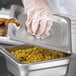 A person in gloves holding a Carlisle stainless steel slotted hotel pan cover over a container of corn.