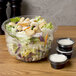 A salad in a Dart clear plastic bowl next to small plastic cups filled with dipping sauces.