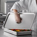 A person wearing a white coat and plastic gloves opening a Carlisle stainless steel steam table pan lid to reveal vegetables inside.