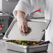 A chef in a white coat holding a Carlisle stainless steel slotted pan lid over a metal container of vegetables.