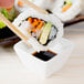 A sushi roll being dipped into a white square porcelain sauce cup.