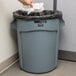A hand throwing a white bag into a Rubbermaid grey trash can.