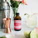 A Nielsen-Massey bottle of rose water on a counter next to a copper cup and a glass of rosemary lemonade.