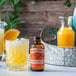 A glass of a drink with orange slices on the rim next to a bottle of Nielsen-Massey Orange Blossom Water.