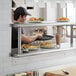 A man in a black hat serving food on a counter with Regency Stainless Steel Pass-Through Shelving above.