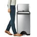 The lower half of a person in jeans standing next to a simplehuman stainless steel rectangular front step-on trash can.
