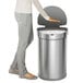 The lower half of a person standing next to a simplehuman brushed stainless steel semi-round motion sensor trash can.