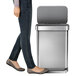 A woman walking next to a simplehuman stainless steel rectangular step-on trash can.