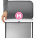A hand inserting a white trash bag with a pink circle and white letter m into a metal trash can.