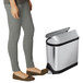 The lower half of a person standing next to a simplehuman brushed stainless steel rectangular trash can.